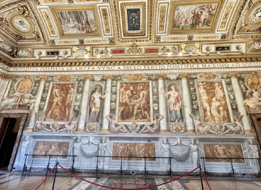 An empty, ornate hall in Castel Sant'Angelo with elaborate frescoes and a gilded ceiling, bathed in warm light